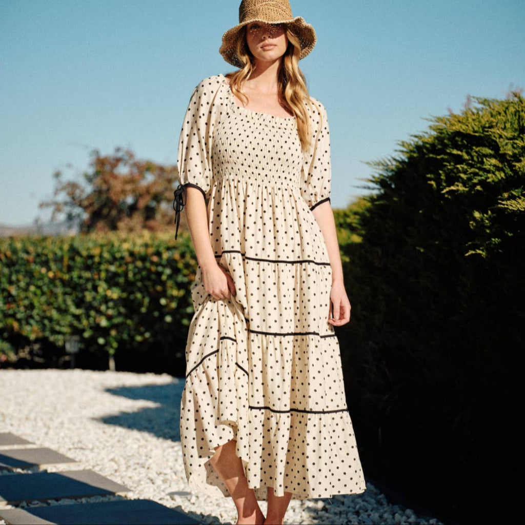 This maxi dress features a timeless polka dot print on a lightweight, breathable fabric. The smocked bodice ensures a comfortable yet flattering fit, while the 3/4 sleeves with adjustable tie details add a touch of elegance. The tiered skirt, accented with contrasting black trim, creates graceful movement.