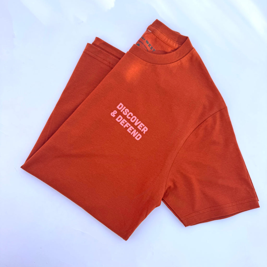 Hand-drawn graphics in faded blush and vibrant pink pop against the fabric's warm, earthy orange tone. A simple, bold front states the mission, "Discover &amp; Defend." The back features a checklist of federally-managed parks in Utah, surrounded by desert flora, fauna, and landmarks.