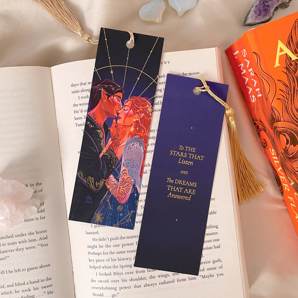 Feyre and Rhysand Gold Foiled Bookmark featuring a gold foiled quote on the back with a hand-tied gold tassel. The front of the bookmark shows two people in a loving embrace wearing deep blue clothing adorned with constellations. The back of the bookmark says, "To the stars that listen and the dreams that are answered". 