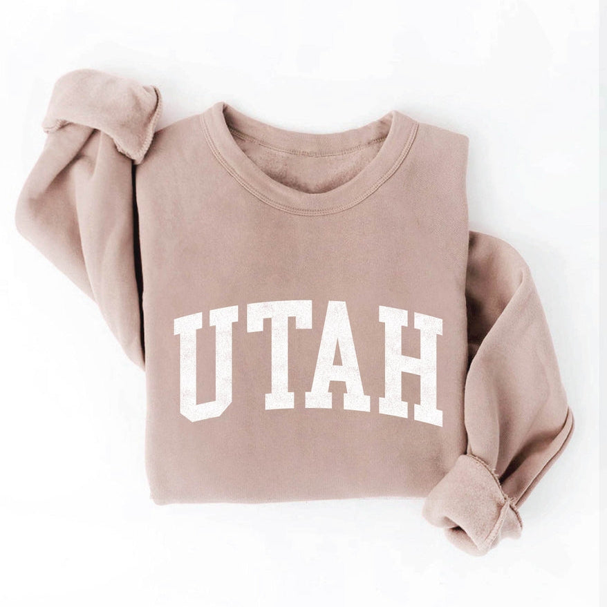 Get the "must-have" look with our Utah Graphic Sweatshirt! This soft, sponge fleece fabric in a beautiful tan color is the perfect unisex pullover. The ribbed cuffs and waistband add a touch of style to this crewneck, featuring "Utah" in eye-catching font. This is perfect for your next ski trip, throwing over your hiking clothes or strolling the streets of Salt Lake City.