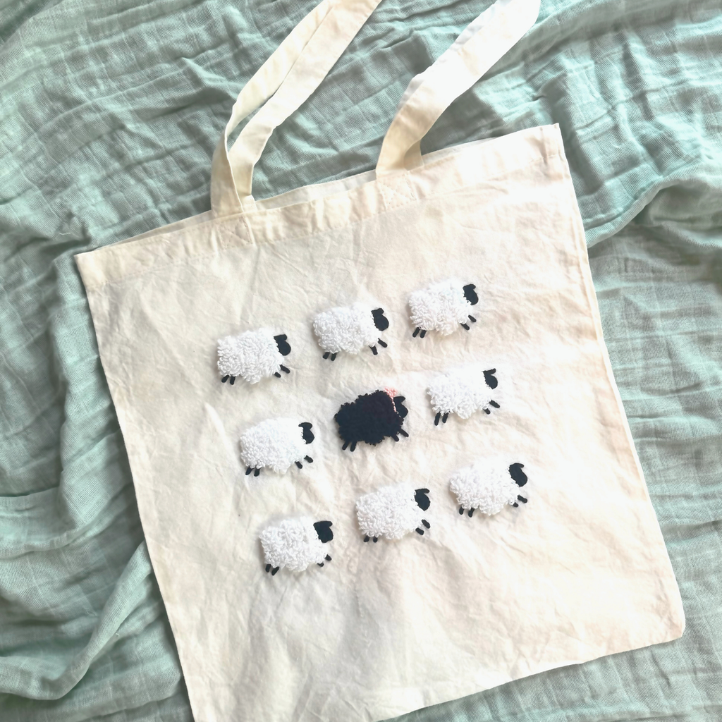 cream tote bag with 9 sheep. the middle 1 is black and the surrounding 8 are white