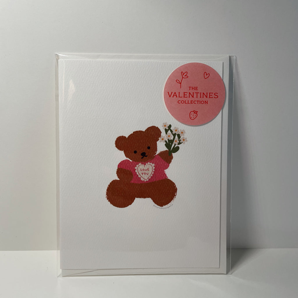 white valentine's day card with a teddy bear wearing a pink shirt that says "love you" and holding a bouquet of white flowers