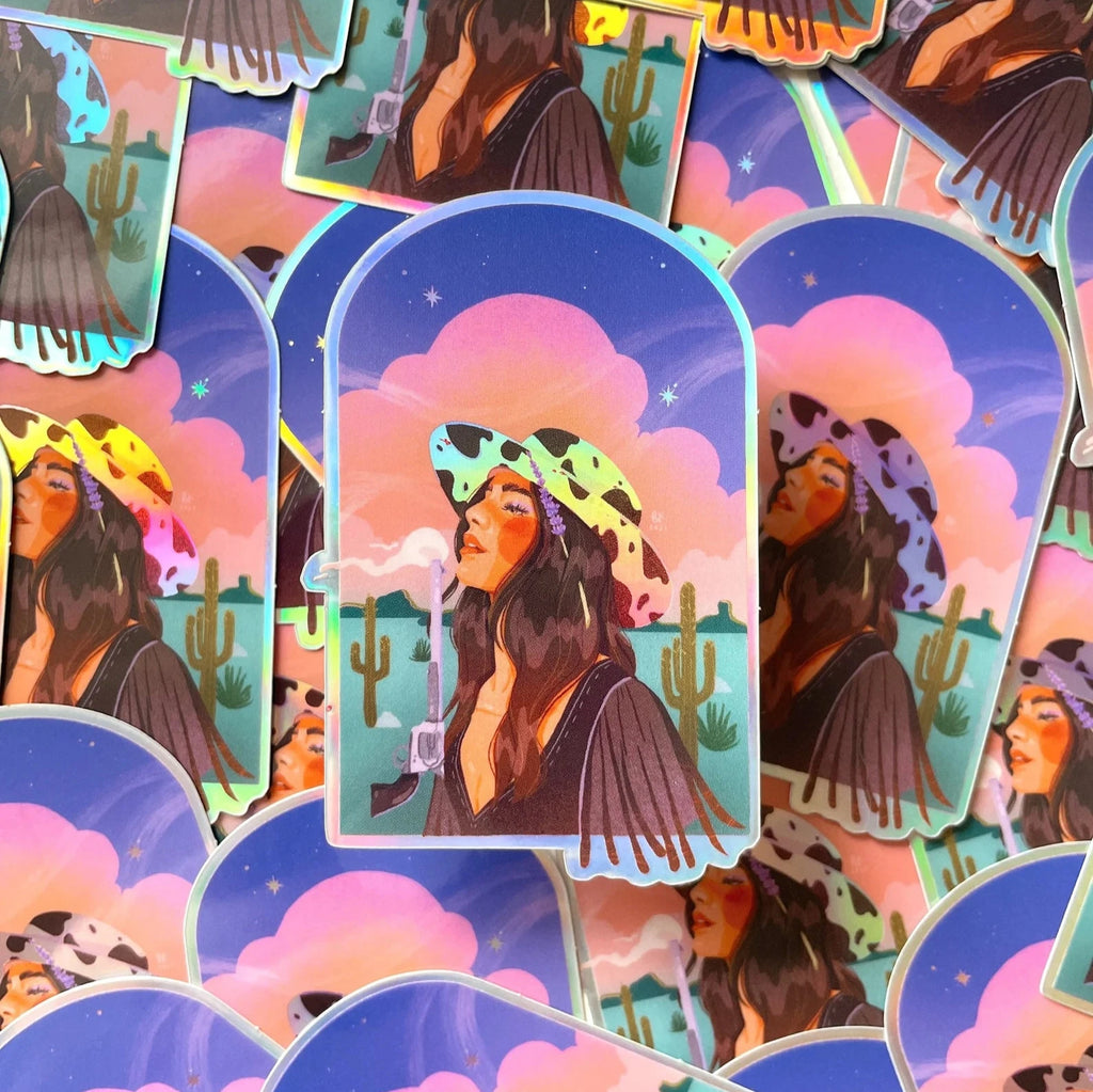 This sticker has a glossy vinyl finish with holographic elements in her hat and on the cutline. A cowgirl wearing a cowprint cowgirl hat has a smoking gun pointed to the sky. She has long brown hair and is facing west. She is in a desert with cacti behind her and a sky with pink clouds. 