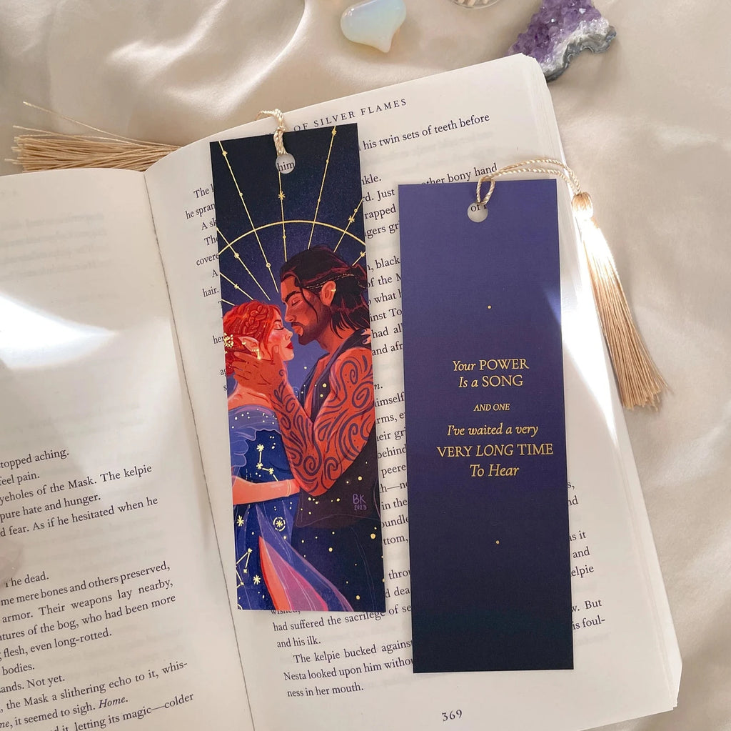 A bookmark with art and a gold tassel attached. On the front of the bookmark is artwork of two people in a loving embrace. one of them is wearing a blue dress adorned with constellations. The other has a muscly arm with tattoos. The back of the bookmark says, "Your power is a song and one I've waited a very long time to hear". 