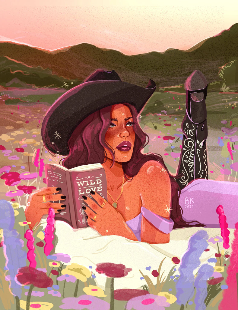 A cowgirl with long brown hair wearing a cowgirl hat and detailed cowgirl boots lies on her stomach on a blanket in a flowery field. She is reading a book titled "wild love"