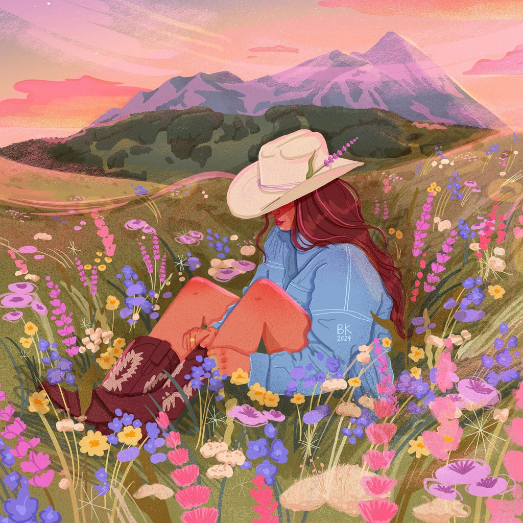 A cowgirl is sitting in a field of botanicals with a large mountain range behind her. She has an ivory cowgirl hat with a piece of lavender sticking out in place of a feather. She is sitting with her knees propped up as she adjusts her boots. 