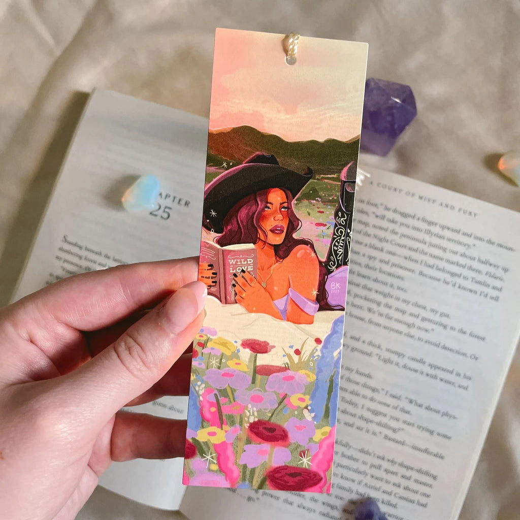 A bookmark with a golden hand-tied tassel. An illustration of a cowgirl wearing a cowgirl hat and detailed cowgirl boots. She is laying on her stomach on a blanket in a flowery field while reading a book titled "wild love"