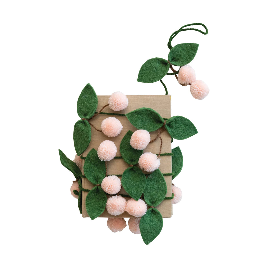 This handmade wool felt leaves and pom pom cherries garland is a whimsical creation that captures the essence of a verdant orchard. Crafted with care, each leaf and cherry pom pom is made from multicolored wool, creating a playful contrast of green, pink, and brown.